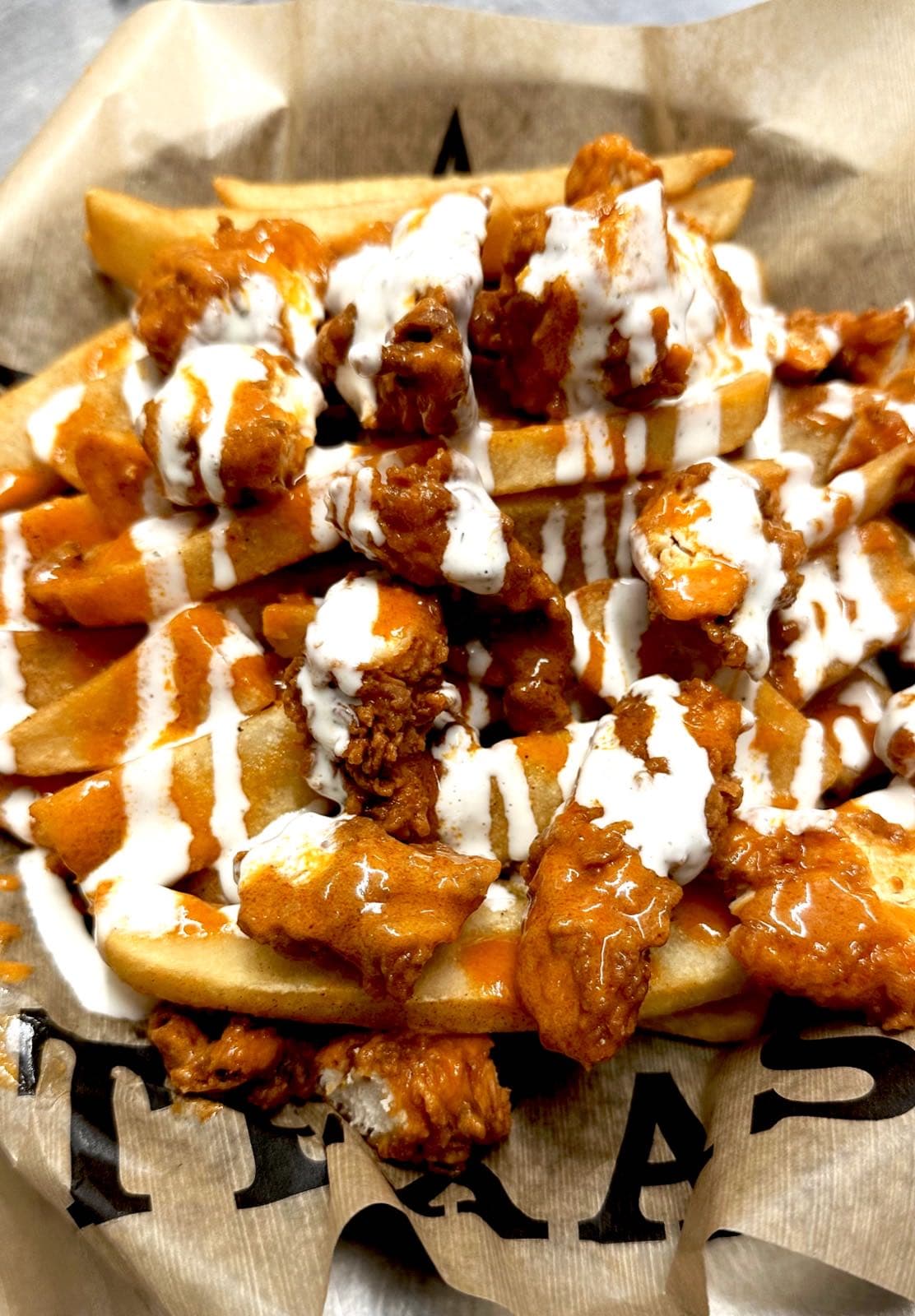 Basket of crispy fries piled with fried chicken bites, drizzled with buffalo sauce and ranch, in a Texas Traditions branded paper liner.