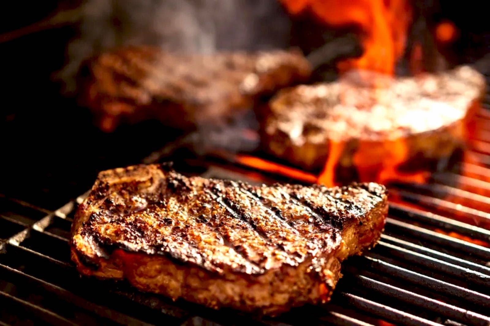 Thick-cut ribeye steaks with dark grill marks searing over open flames on the grill grate.