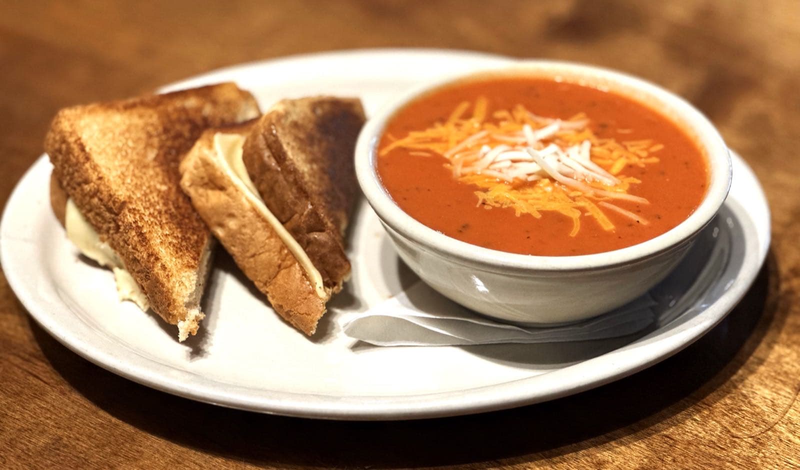 A toasted grilled cheese sandwich cut in half beside a bowl of tomato soup topped with shredded cheddar and Monterey Jack.